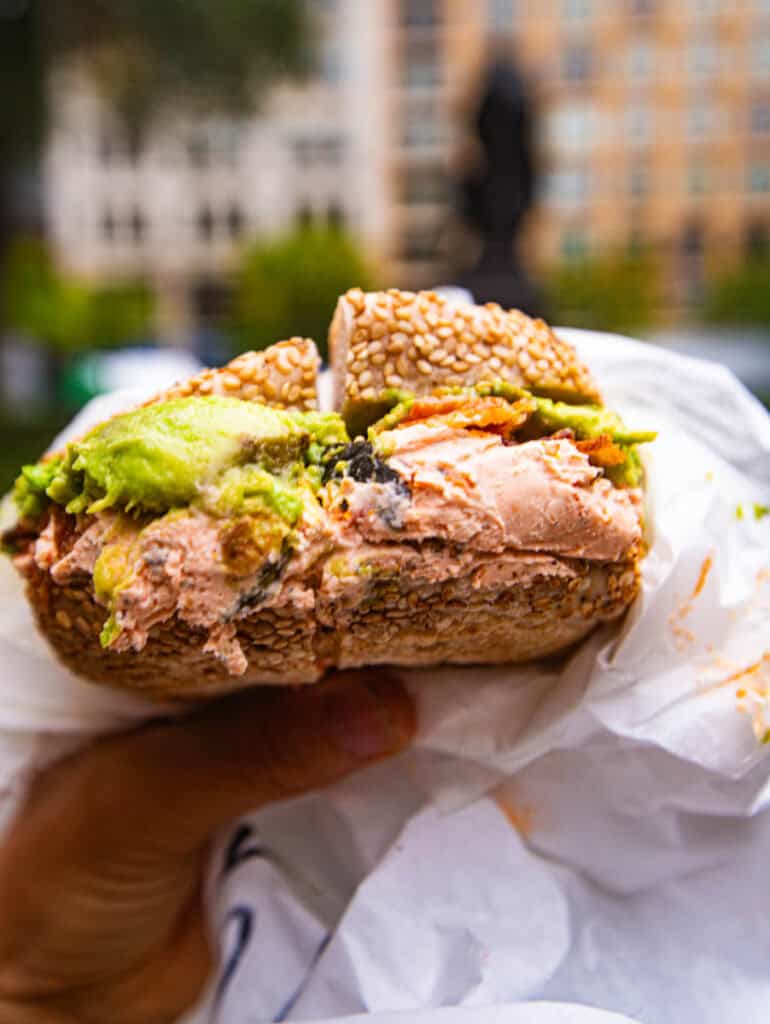 person holding bacon avocado sun dried tomato cream cheese bagel