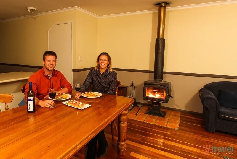 man and woman sitting at table by the fire with plates of food and red wine. 